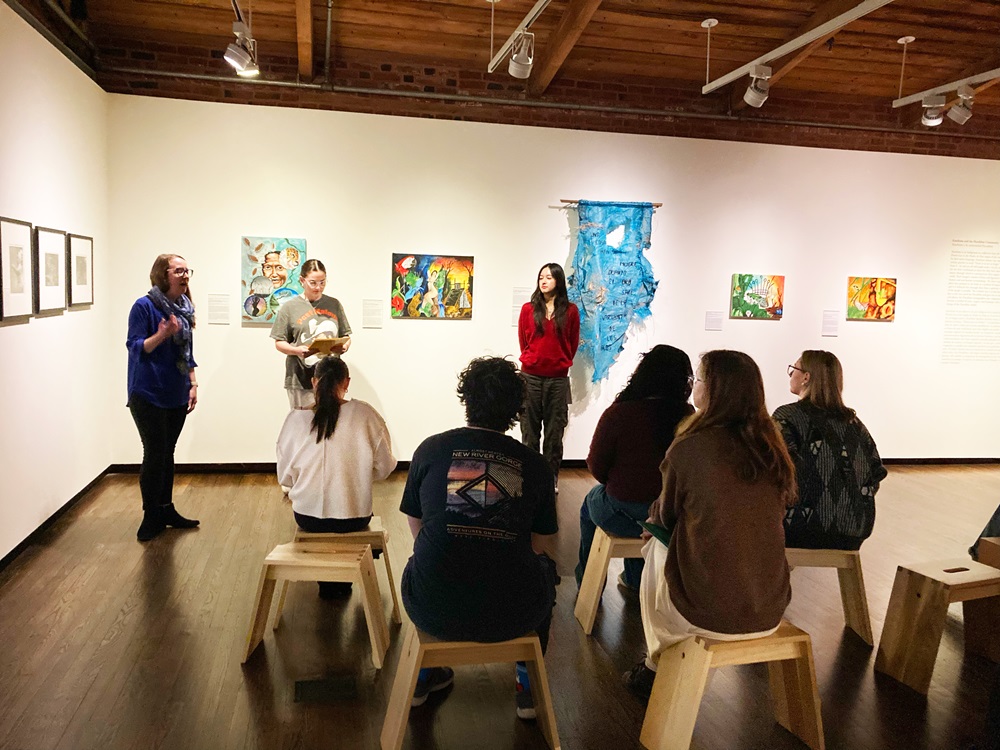 Three people stand along a wall in a gallery covered in art, speaking to a seated group of listeners.