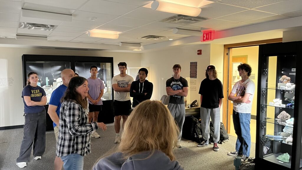 Class session in a gallery with cases of minerals. Woman addresses a group of students.