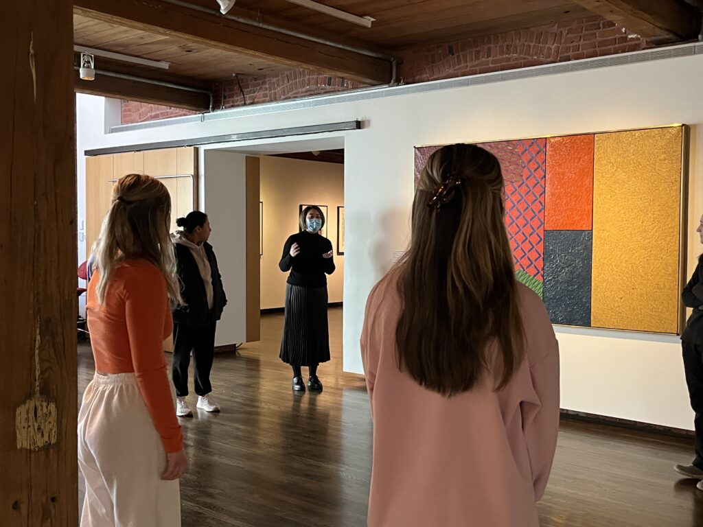 Instructor teaching a class in art gallery. Group standing near large, colorful, abstract painting.