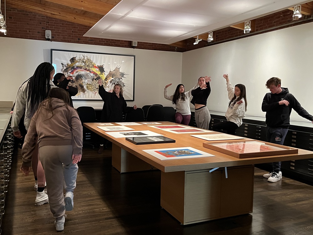 Group of students standing around a large table with art laid upon it. Students are making dance poses.