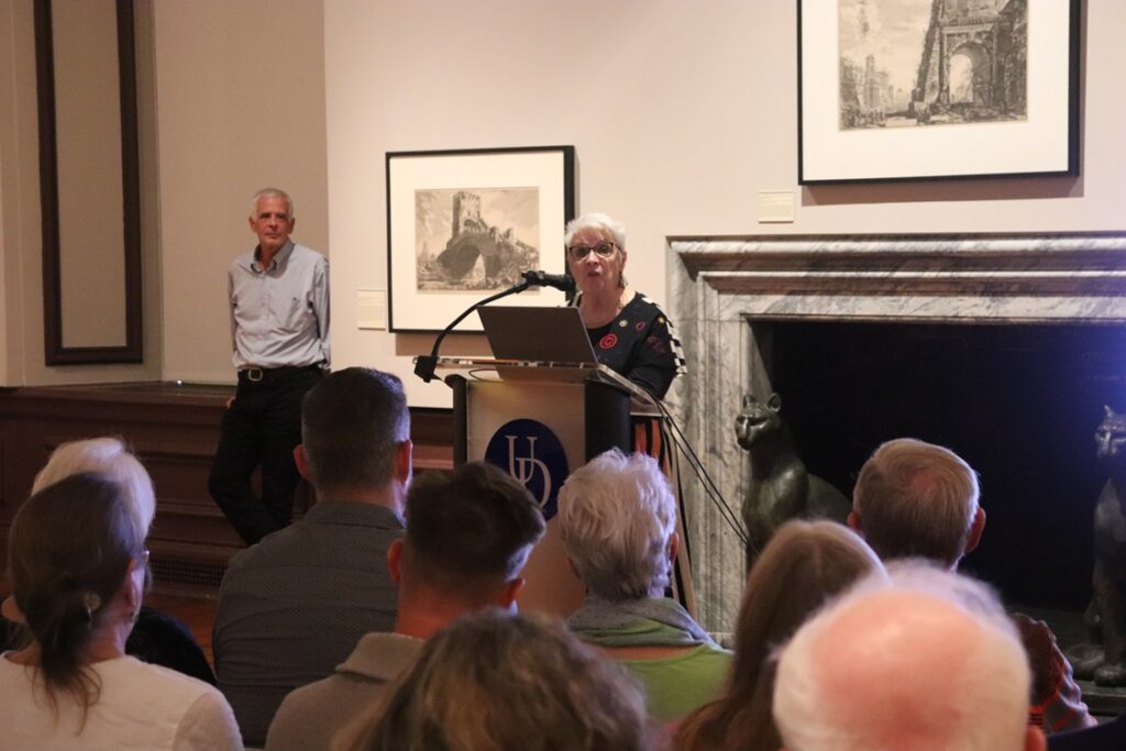 Woman at podium in front of a fireplace lecturing to a seated group of visitors in an art gallery. One man stands behind the speaker to the left