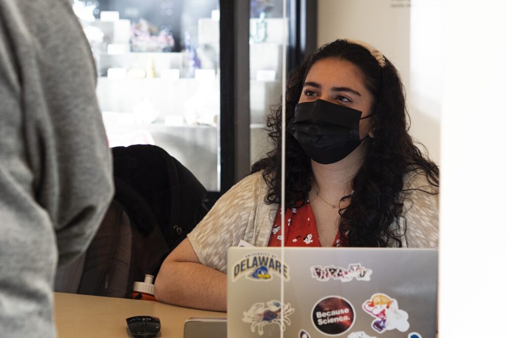 Woman sitting at desk in front of a case of minerals. Laptop open in front of her. She wears a face mask