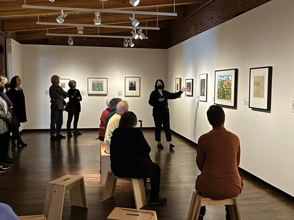 Standing woman gesturing to art on the wall, speaking to a group of visitors in an art gallery