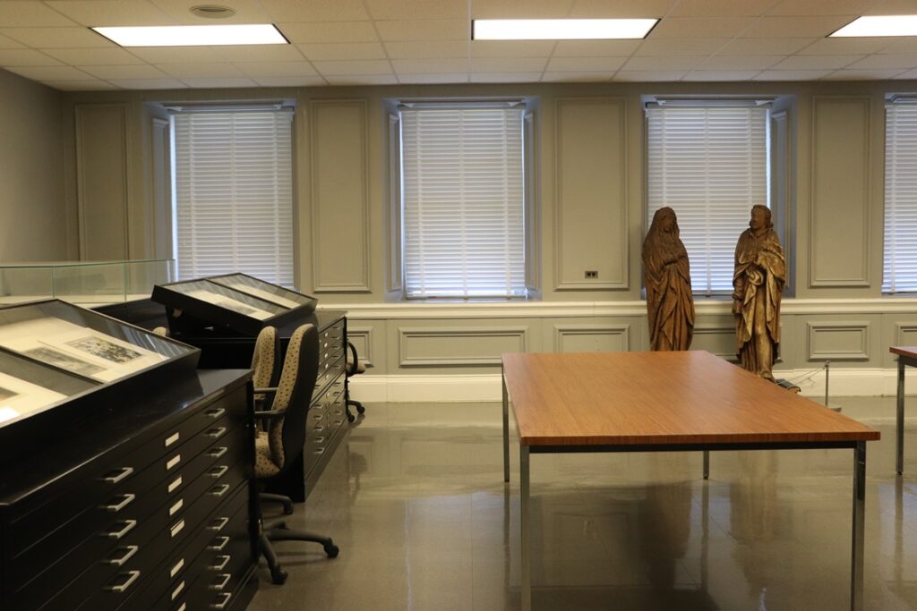 Classroom with empty table in and row of flat files and art in cases to the left. Two sculptures in background in front of a row of windows.