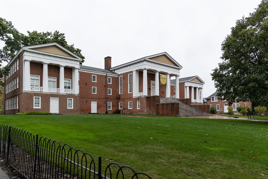 Large, three-story brick building with large central staircase to front door on second floor. Three pediments and white columns. Large lawn in foreground.
