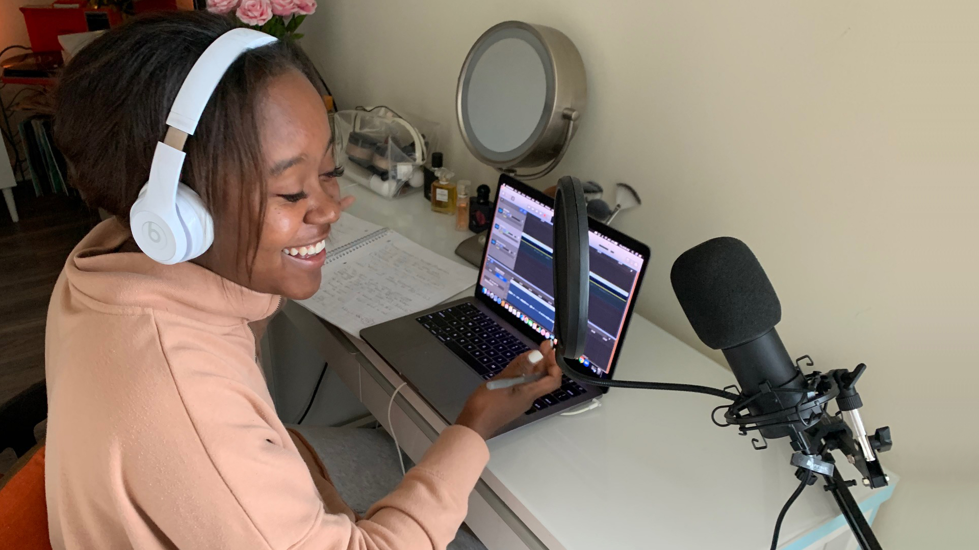 Young woman sitting at a table with her laptop open, while recording a podcast. She has headphones on and is speaking into a microphone.