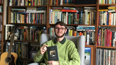 A young man in a green sweater holding a book in his hands as he sits in a chair. He is sitting in front of a massive wall of bookshelves, filled with books stored in a variety of directions.