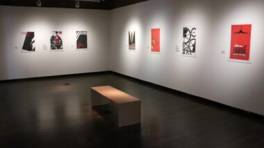 A darkly lit room with a bench in the middle and mounted posters spread out evenly on white walls. The posters are spotlighted with bright lights.