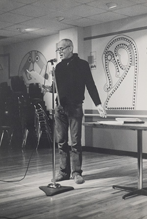 A black and white photo of Edward Field standing at a microphone and reading.