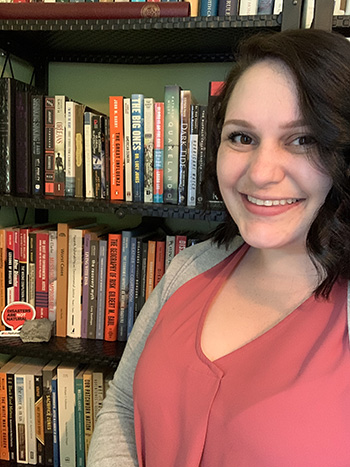 A young woman smiling. She is standing in front of several packed bookshelves.