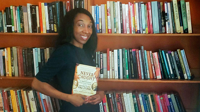 A young woman holding a book and standing in front of several large bookcases that are filled with books.