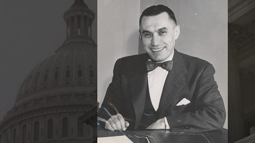 In the foreground, a black and white photograph of a man sitting at a desk with a couple of documents. He is smiling and wearing a suit and bowtie. In the background, an image of the U.S. Capitol.