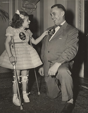 A black and white photograph of a young girl with leg braces and crutches who is pinning a badge on a man kneeling beside her. Both individuals are wearing March of Dimes badges and smiling.