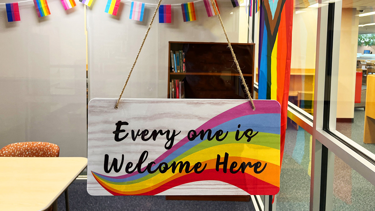A look into a brightly lit room with pride flags, a bookshelf, a table and a sign that reads "Everyone is welcome here: