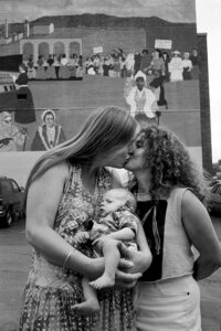A black-and-white photograph of two women kissing and holding a baby between them in front of a mural of women throughout history.