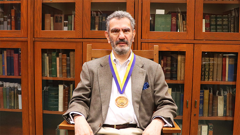 An individual sitting in a chair in front of several shelves of enclosed bookcases. The individual is wearing a medal around their neck.
