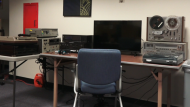 A desk with computer screen, chair and several pieces of equipment to play older forms of media on them