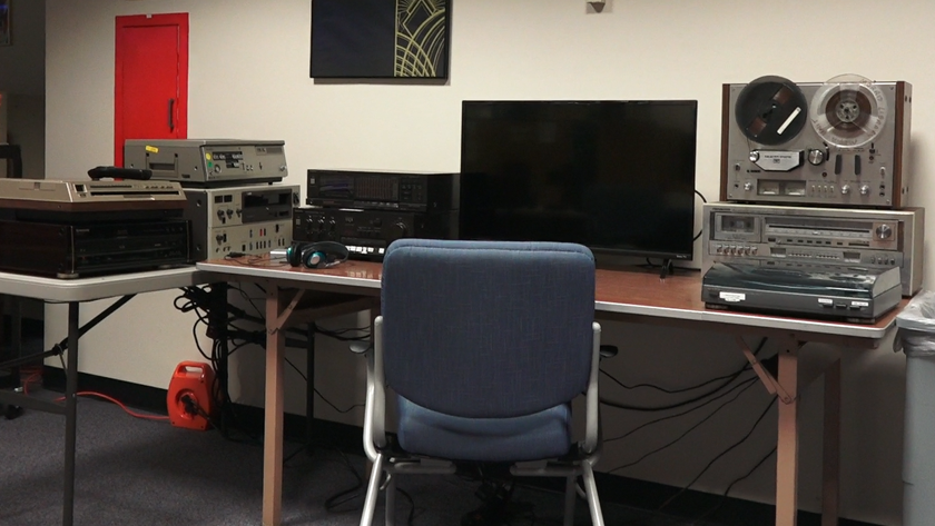 A desk with computer screen, chair and several pieces of equipment to play older forms of media on them