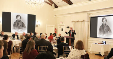 A banquet room with a speaker at a podium and a crowd seated around round dinner tables.