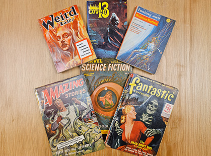 Six bound periodicals with titles like "Weird Tales", "Coven 13" and "Amazing Stories" are arranged on a table top.