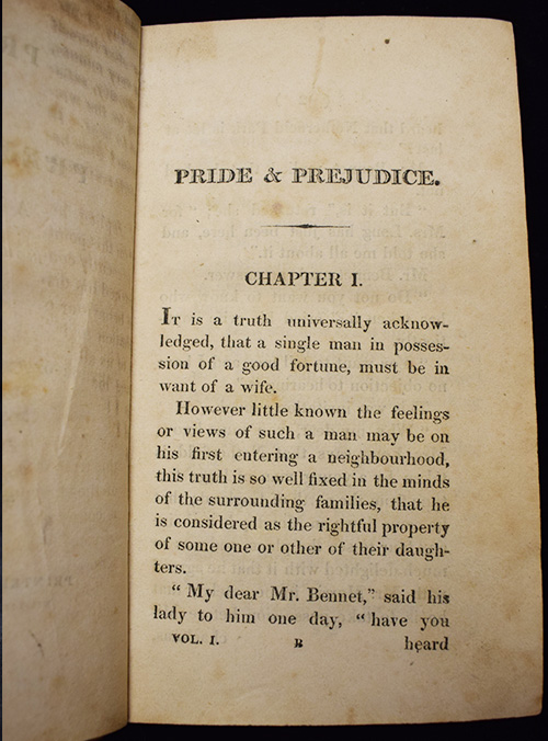 The opened first volume of a first edition of Jane Austen's novel, Pride and Prejudice, showing the beginning of Chapter 1. The pages are yellowed, and the text begins with the famous line: "It is a truth universally acknowledged, that a single man in possession of a good fortune, must be in want of a wife."