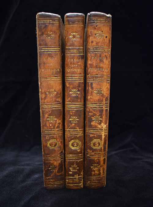 Three leather-bound volumes of the first edition of Jane Austen's novel, Pride and Prejudice, standing vertically against a dark background. The spines are brown leather, stamped with gold decorations of stars and horizontal bands. The title "Pride and Prejudice" is visible in gilt text on each volume's spine, along with the volume number 1, 2, and 3 at the bottom.