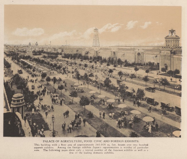A high-angle, sepia aerial photograph overlooking a bustling walkway outside the Palace of Agriculture. The broad thoroughfare is filled with fairgoers strolling between manicured green lawns, young trees, and parked vintage automobiles.