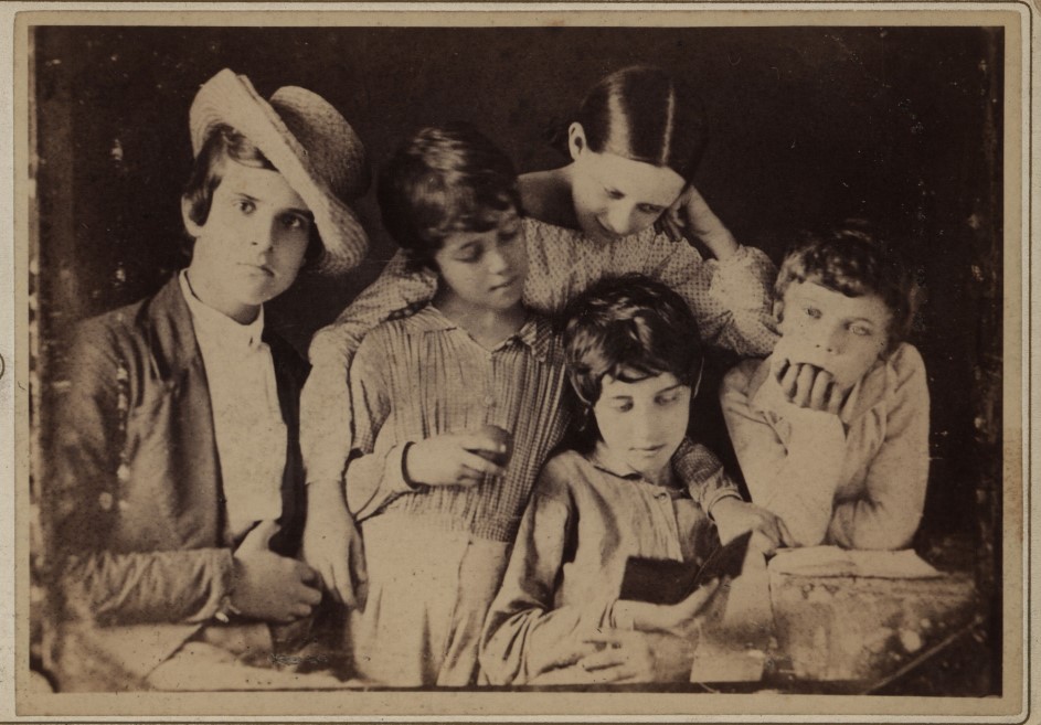 Five children in period clothing crowded around a small book, one reading while the others watch with curiosity—two leaning in, one resting chin on hand, and another wearing a straw hat.
