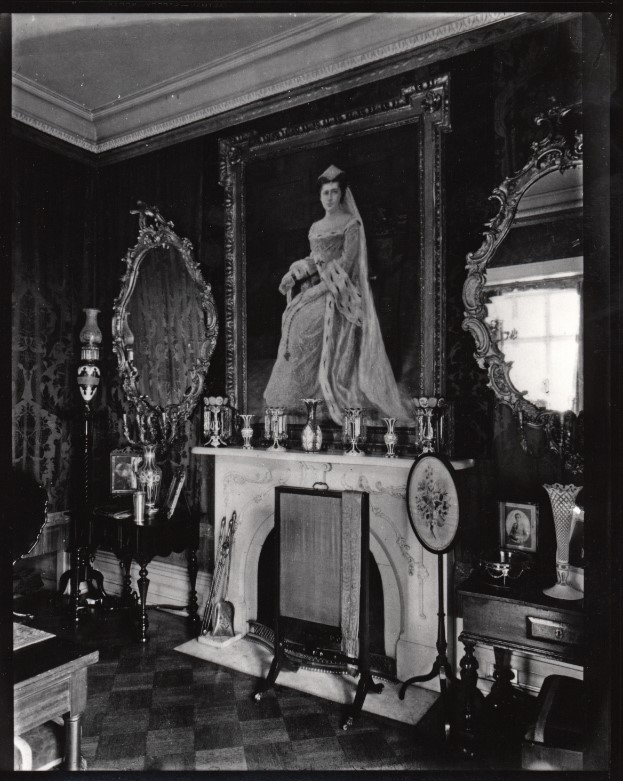Ornate Victorian parlor centered on a marble fireplace topped by a large formal portrait of a woman in a gown, flanked by two carved mirrors and a display of silver trophies, urns, and decorative objects indicating 19th-century domestic wealth and taste.