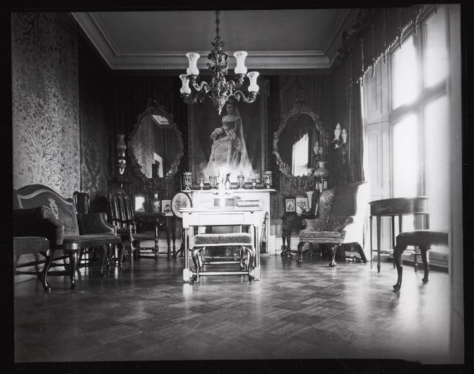 Victorian-era formal parlor arranged for receiving guests, featuring a central fireplace topped by a large portrait, an ornate chandelier, two carved mirrors, grouped antique chairs and small tables, and tall windows casting light across a parquet floor.