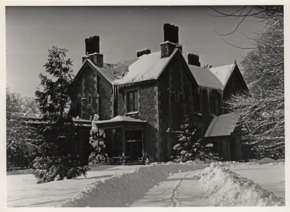 Historic stone house with steep gables and tall chimneys blanketed in snow, icicles hanging from the eaves; a shoveled path with faint footprints leads to the front door amid snow‑laden evergreen trees, conveying a quiet winter scene.