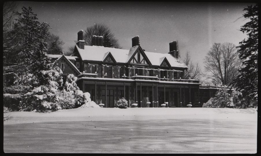 Victorian Tudor-style mansion with steep gables, tall chimneys and a wide front porch, set in a quiet winter scene with heavy snow on the roof, trees and a frozen lawn in the foreground, evoking a cold, serene atmosphere.