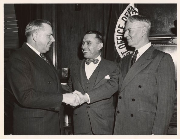 A black-and-white photograph depicting three men in formal suits standing together indoors. The two men on the left are shaking hands and smiling, while the man on the right, wearing glasses, stands alongside them. A Post Office Department seal is partially visible on the wall behind them.