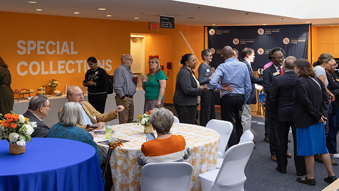 Guests mingle and talk in small groups around tables at a reception in front of a University of Delaware Special Collections wall.