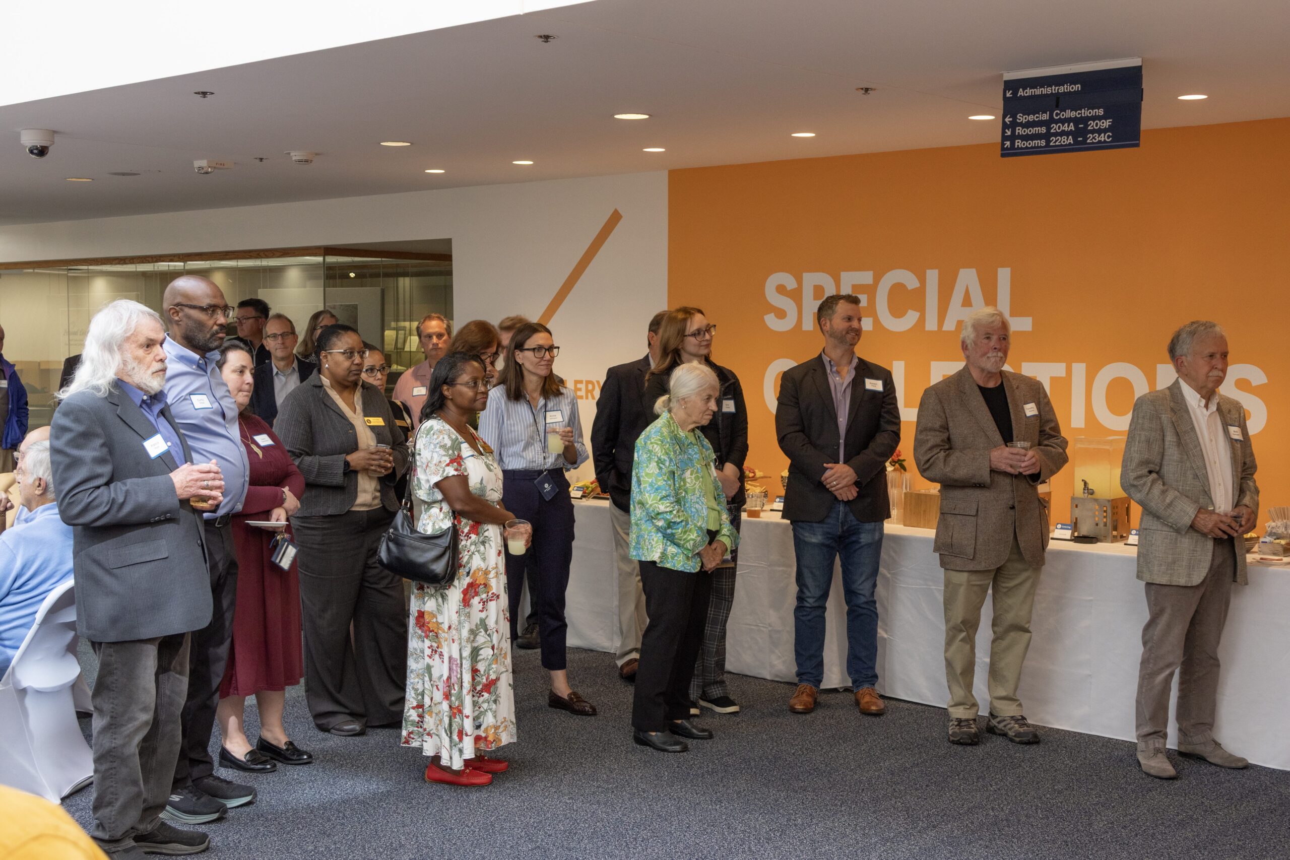 Attendees socializing in small groups and seated around decorated round tables at an indoor reception, with a bright orange "Special Collections" wall and a University of Delaware backdrop in the background.