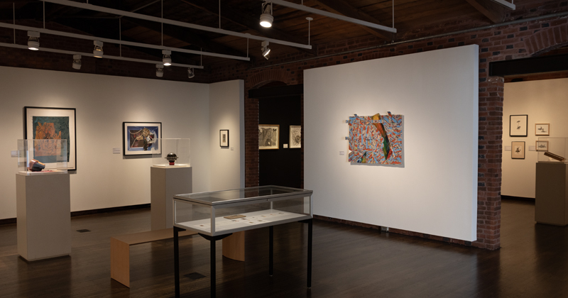 An art gallery interior with dark hardwood floors, exposed brick pillars, and overhead track lighting. The room features framed abstract artwork on white walls, small sculptures displayed under glass on pedestals, and a central glass display table.