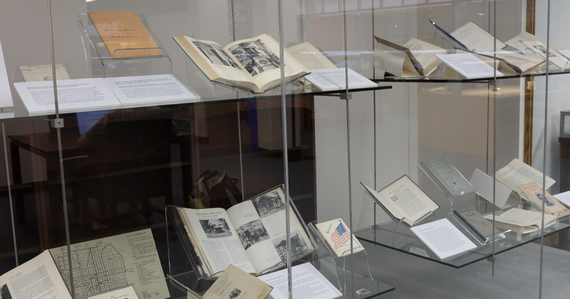 A multi-tiered clear glass display case filled with open historical books, pamphlets, architectural maps, an American flag card, and explanatory text placards.