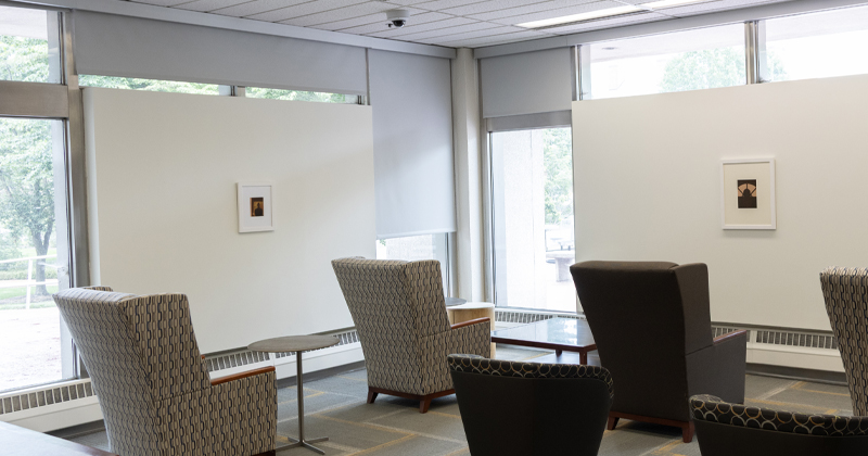 A brightly lit gallery seating area with patterned armchairs, small tables, and large windows, displaying two small framed artworks mounted on white partition walls.