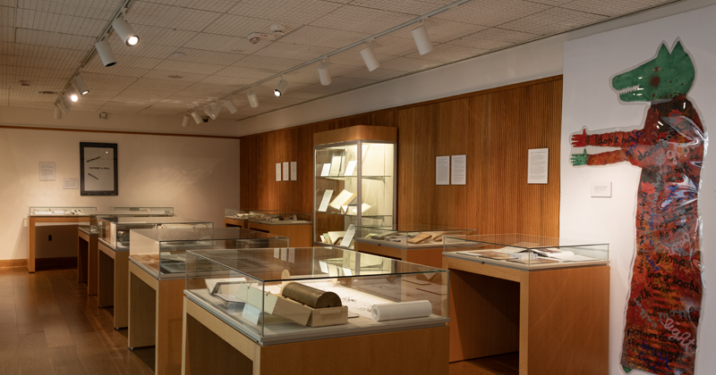 An exhibition hall with wood-paneled walls and glass cases filled with books and archival documents, featuring a large cut-out artwork of a green-headed figure in a patterned red coat on the right wall.