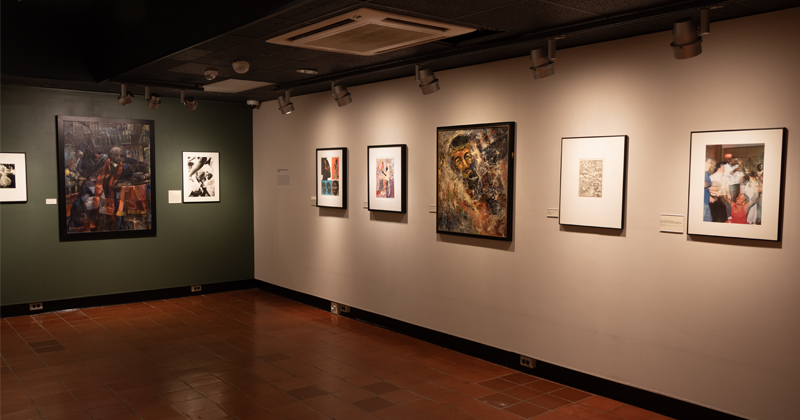 An art gallery room illuminated by track lighting, featuring framed paintings, prints, and mixed-media artwork mounted along dark green and off-white walls.