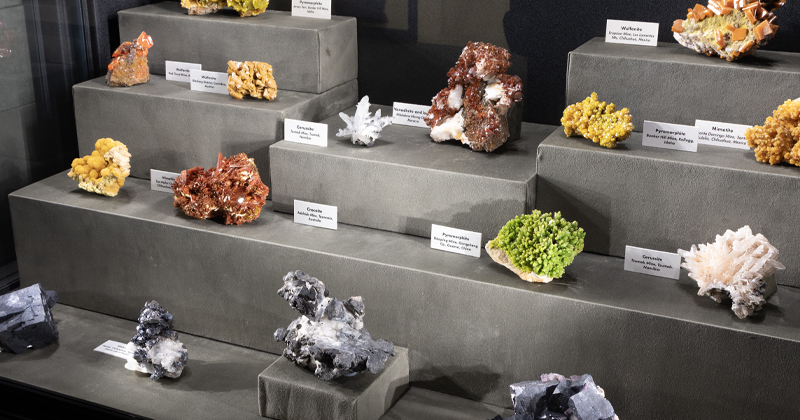 A multi-tiered museum display of colorful mineral specimens including yellow, orange, green, brown, and white crystal formations, each accompanied by small white label cards.