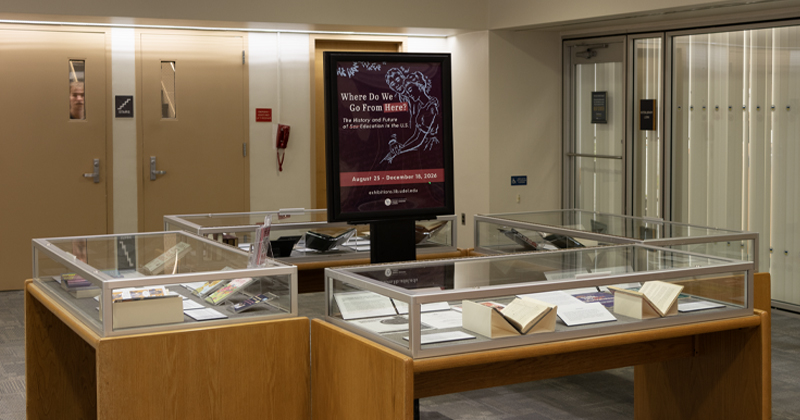 An exhibition room with glass display cases containing books and documents, centered around a digital display screen titled "Where Do We Go From Here? The History and Future of Sex Education in the U.S."