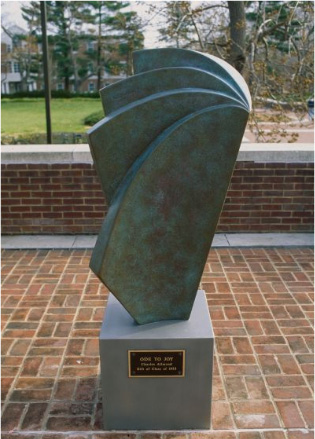 An outdoor photograph of an abstract bronze sculpture with a green patina, titled "Ode to Joy." The artwork consists of overlapping, fan-like curved geometric plates mounted on a light gray pedestal with a brass plaque, situated on a brick courtyard with trees and a building in the background.