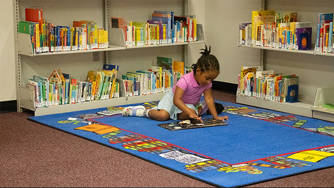A young girl sitting on a colorful children's rug in a library corner, focused on reading a picture book while low shelves of children's books surround her.