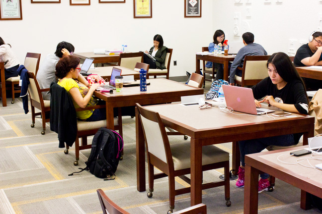 Students quietly studying at wooden tables in a campus library, several using laptops, notebooks, and water bottles while focused on their work.