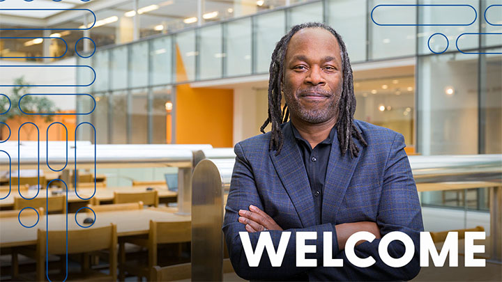 Smiling man with shoulder-length locks wearing a blazer stands in a bright campus library atrium, conveying a warm welcome to visitors; bold "WELCOME" text is overlaid at the lower right.