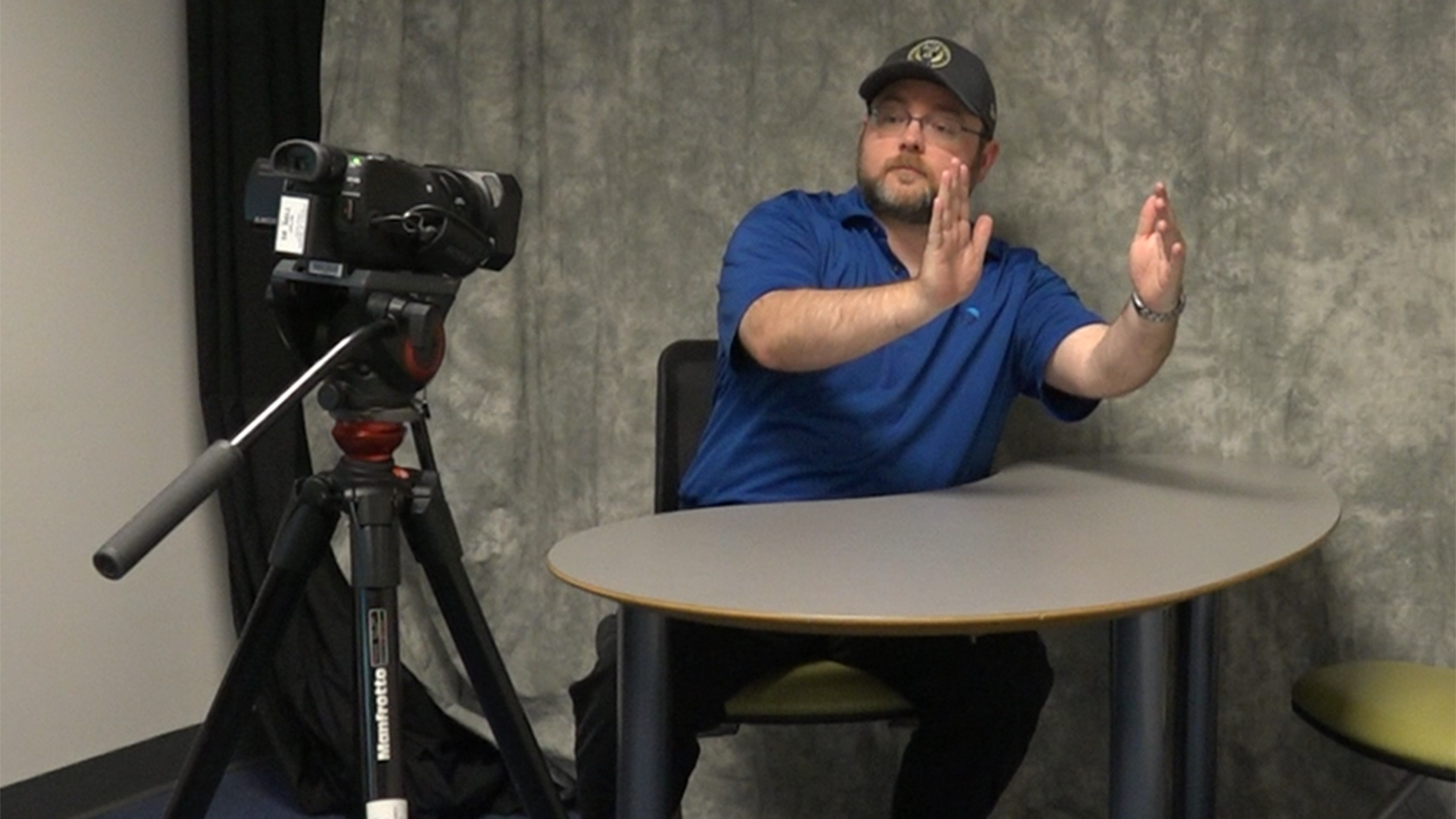 A person sitting at a table in front of a camera. They are using their hands to indicate positioning for the camera.