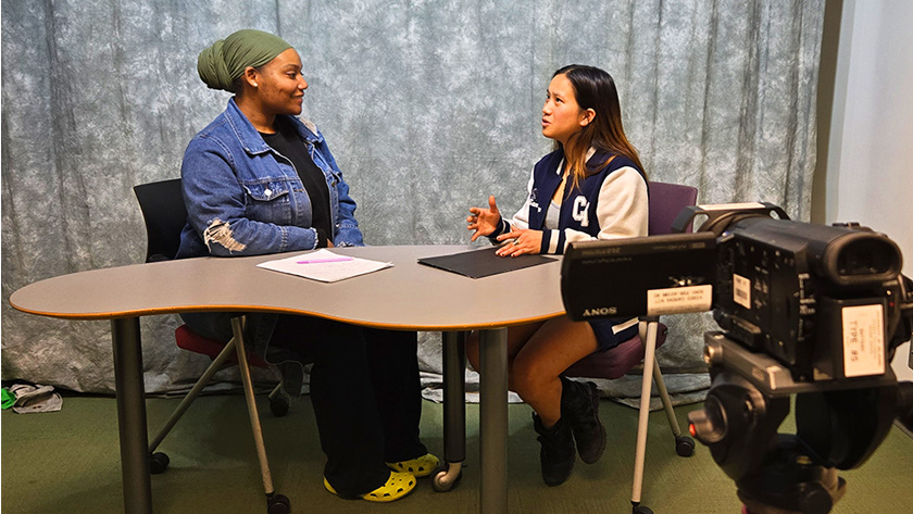 Students sitting and facing each other at a table. They are talking and being recorded by a visible camera in the foreground.